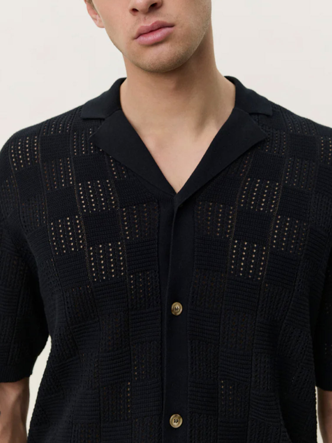Someone wears the Les Deux Squared Knitted SS Shirt, a black short-sleeved button-up with a checkerboard texture and camp collar, shown from shoulders to mid-torso against a plain background.