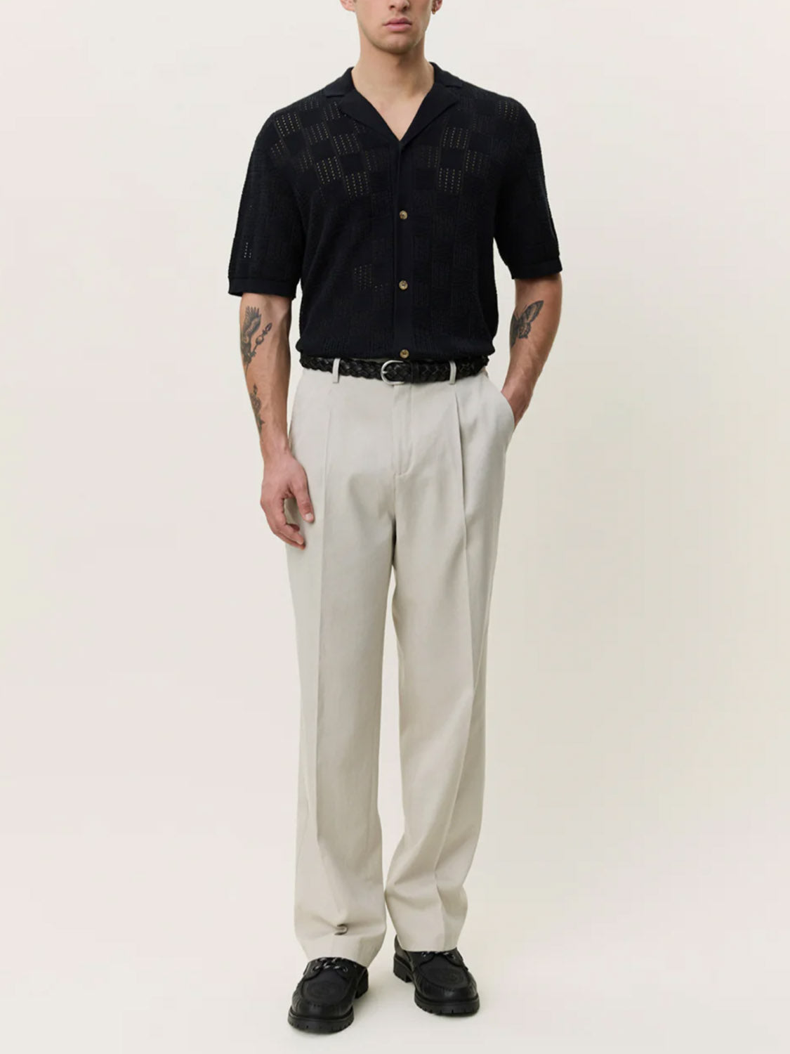 A man with tattooed forearms stands against a plain background wearing Les Deux's Squared Knitted SS Shirt, a black short-sleeve camp-collar top, paired with light beige pleated trousers, a black belt, and black shoes. His hands are in his pockets.