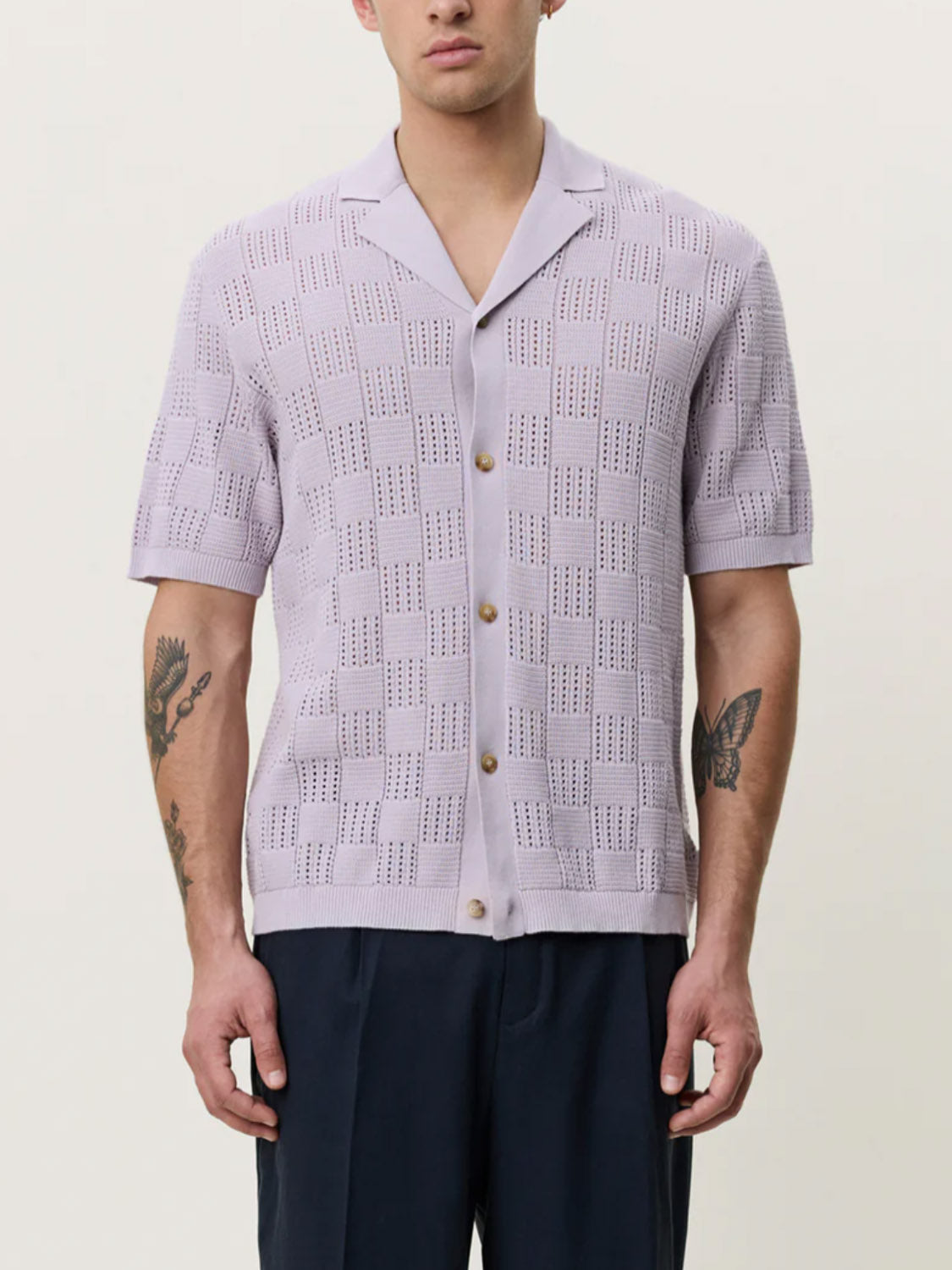 A person wears the Les Deux Squared Knitted SS Shirt—a light purple, short-sleeve cotton knit with a camp collar and geometric perforations—paired with dark pants, showing tattoos on both forearms.