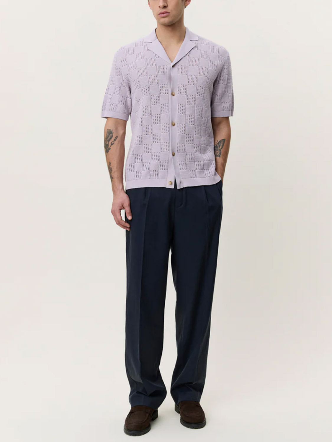 A man wears the Les Deux Squared Knitted SS Shirt, a light purple cotton camp collar shirt, paired with dark loose trousers and brown shoes. His tattooed forearms are visible as he stands with hands in pockets against a plain, light background.