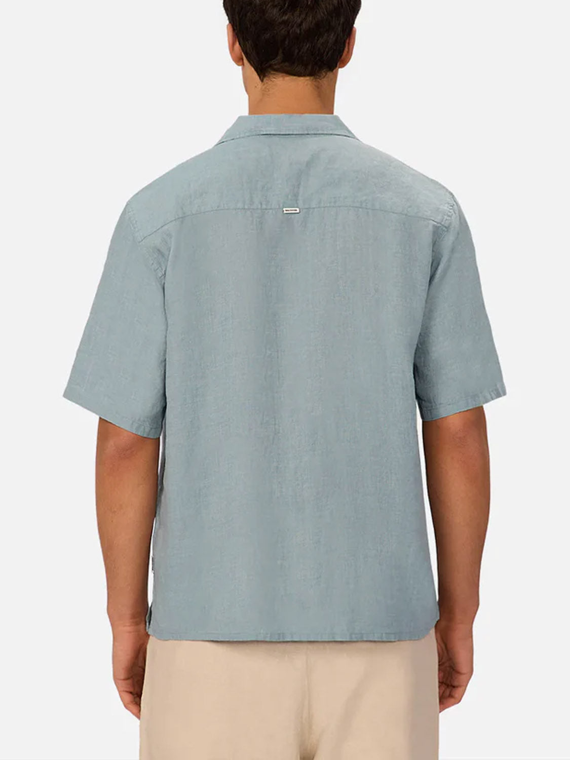 A man is seen from behind wearing Industrie’s The St Martins Short Sleeve Shirt in light blue with beige shorts, set against a plain light background.
