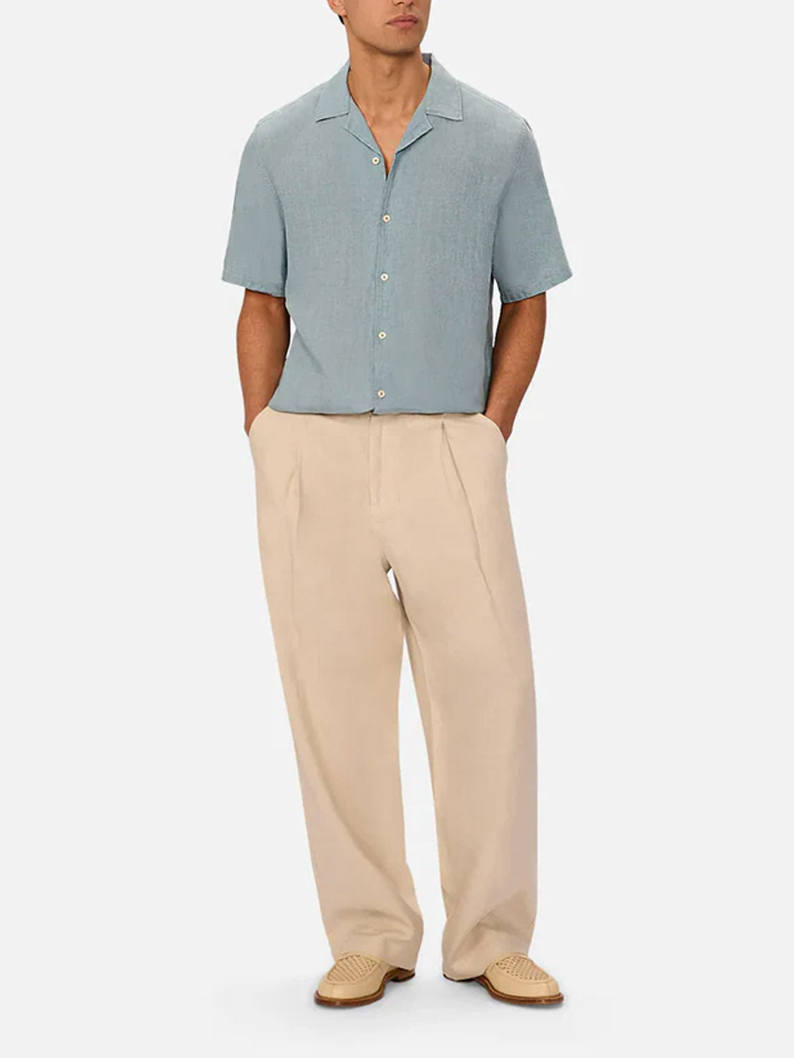 Wearing Industrie’s The St Martins Short Sleeve Shirt in light blue, beige pleated pants, and woven beige shoes, a person stands hands-in-pockets against a plain light background.