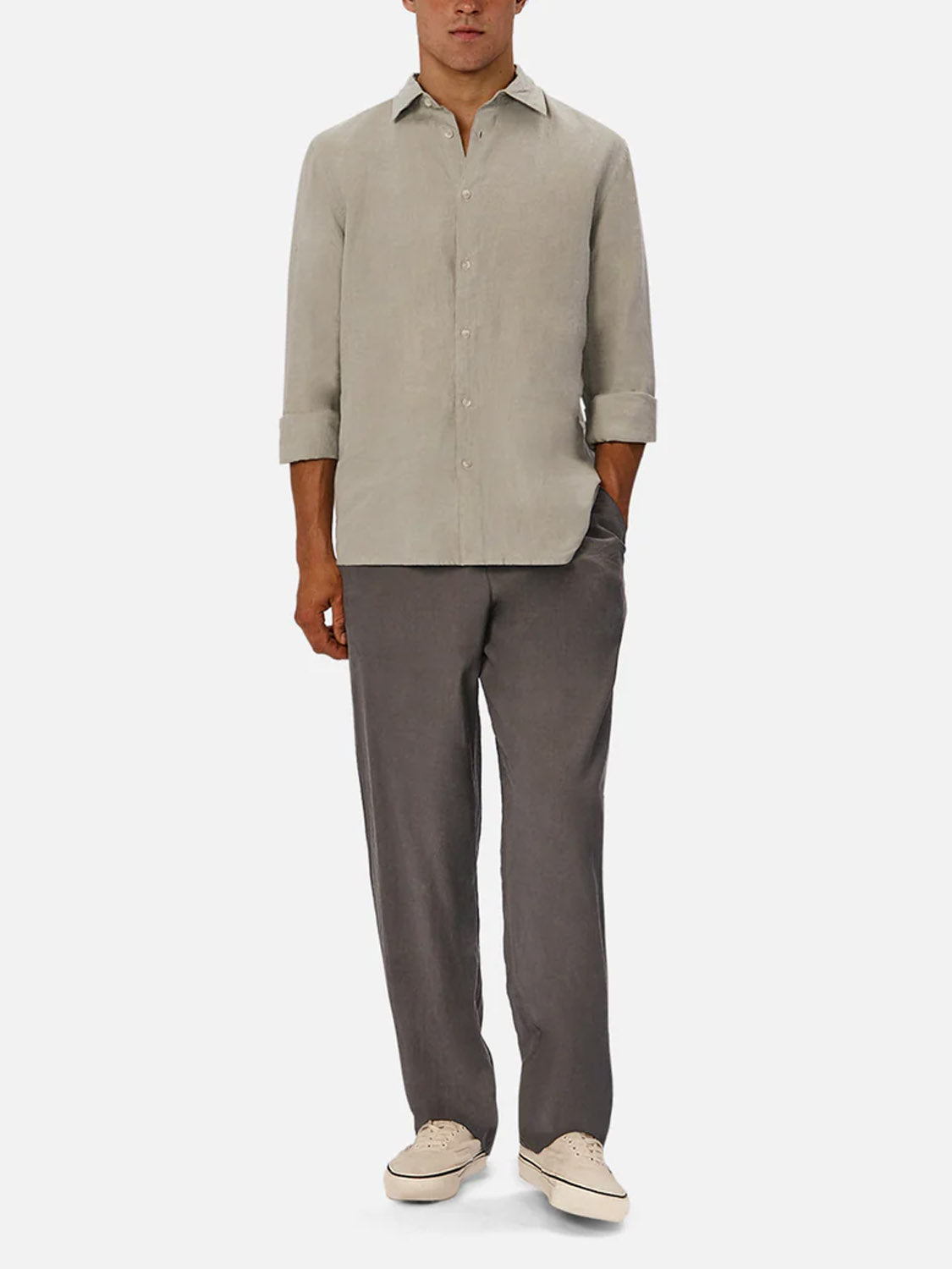 A man stands with hands in pockets, wearing Industrie’s The St Martins Long Sleeve Shirt in light beige, paired with loose gray trousers and beige sneakers, set against a plain white background.