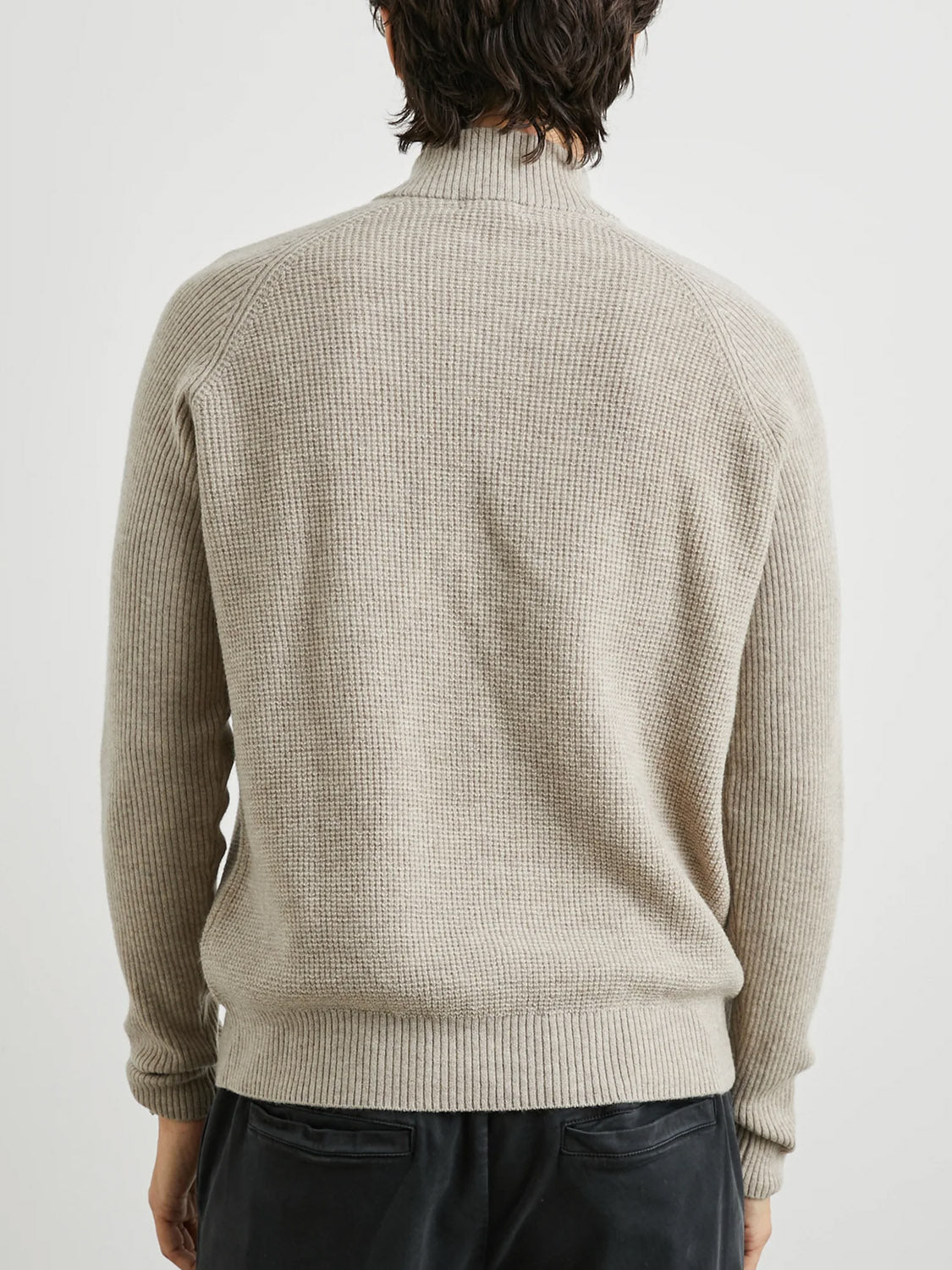 A person is shown from the back wearing the Rails Stark Quarter Zip Sweater, a light beige merino wool piece with long sleeves and a mock neck, paired with dark pants against a white backdrop. Their shoulder-length dark hair complements this versatile layering option for any occasion.