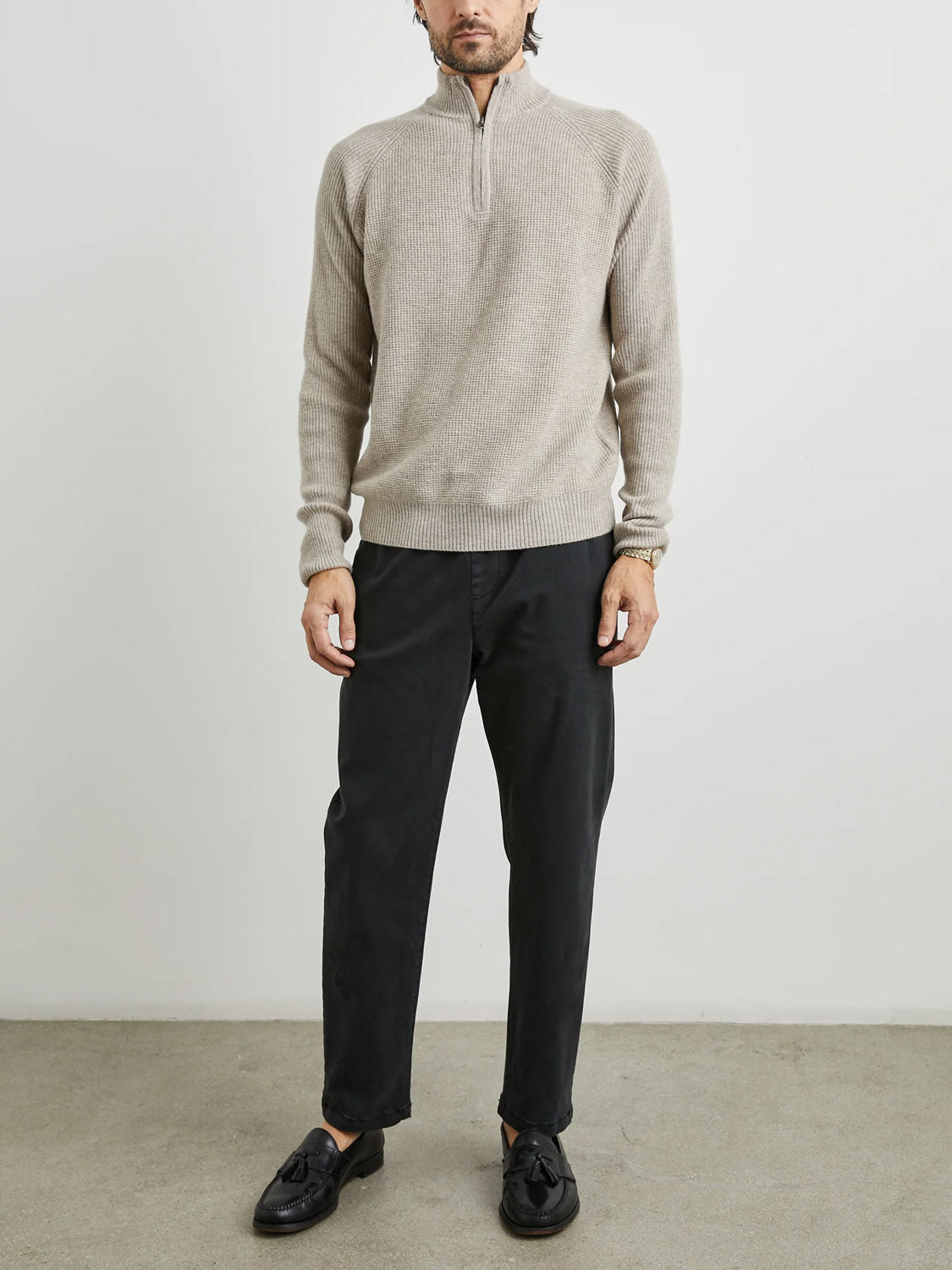 A person stands confidently on a grey surface, wearing a light beige layering piece over the Rails Stark Quarter Zip Sweater in grey, paired with black pants and tassel-adorned black loafers. The plain, light-colored background enhances the ensemble.