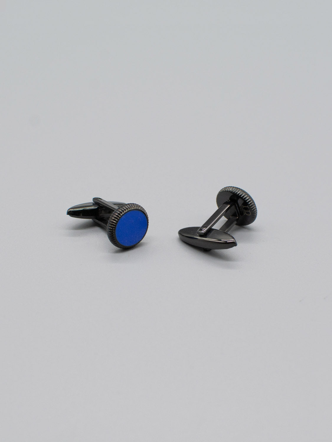 Two Black Gold Plated Steel Enamel Cufflinks by ID Menswear, featuring round blue enamel inlays and a sleek modern design, rest on a gray surface. One stands upright while the other lies on its side, showcasing their polished finish and reliable bullet back mechanism.
