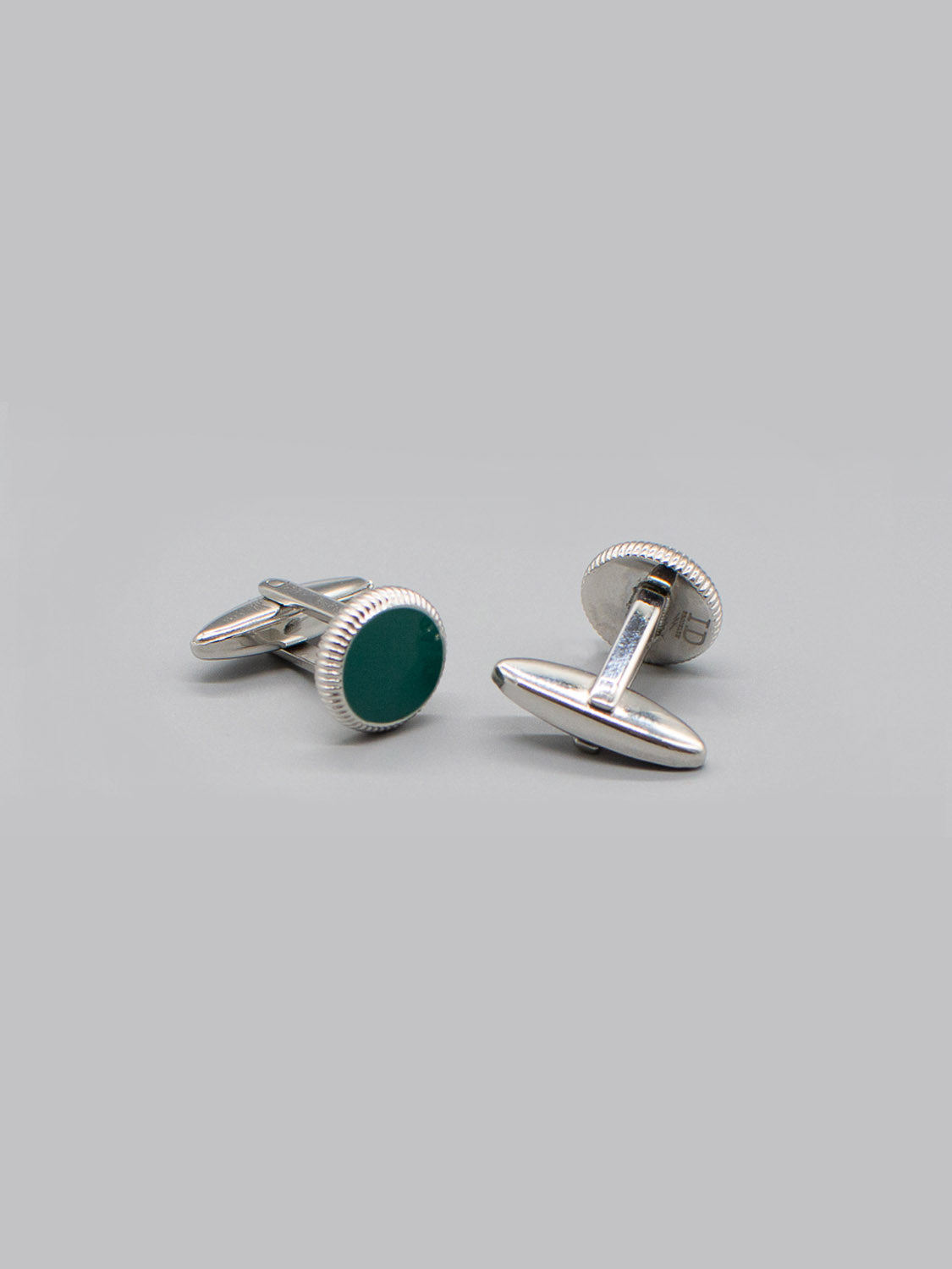 Two Rhodium Plated Steel Enamel Cufflinks by ID Menswear, featuring round green enamel faces, are displayed on a light gray background. One stands upright while the other lies on its side, showing the post and bullet back mechanism.