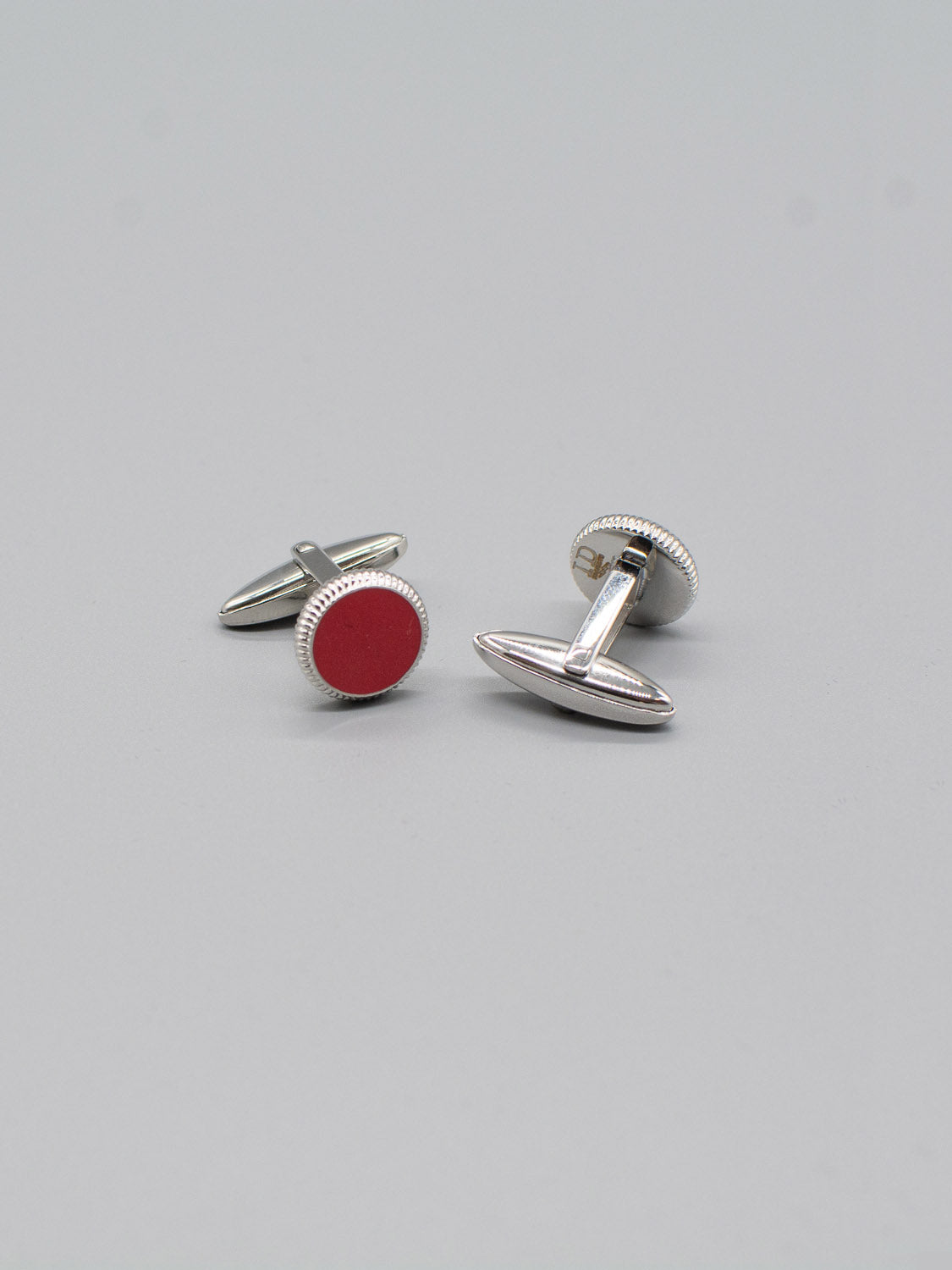 ID Menswears Rhodium Plated Steel Enamel Cufflinks showcase a circular design with a red enamel inlay and silver edges, crafted from stainless steel. Featuring a bullet back mechanism, theyre elegantly displayed on a plain gray background.
