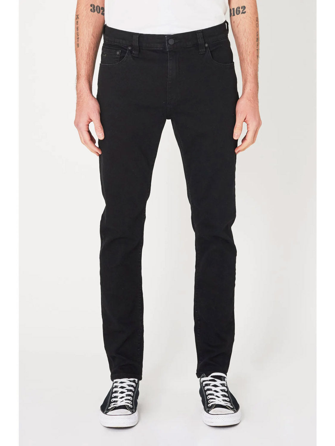 A person wearing Rollas Stinger Black Gold skinny leg jeans, made from super stretch denim, and black-and-white sneakers stands against a white background. The cropped image shows only the lower torso, legs, and tattooed forearms.