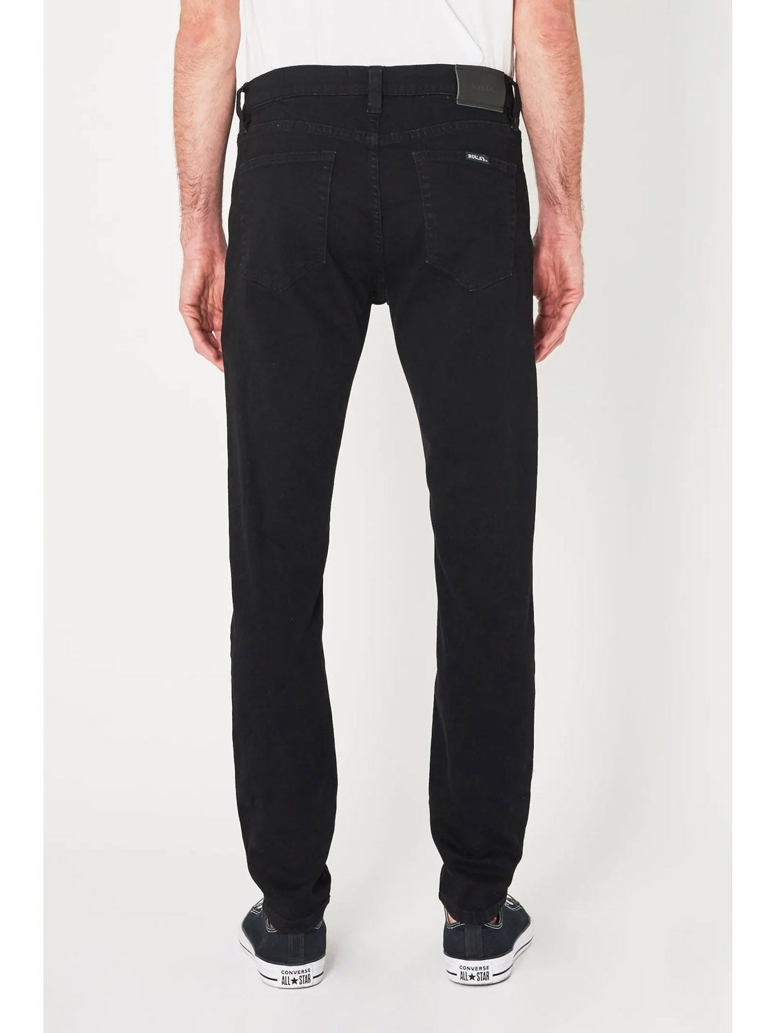 A man faces away from the camera, wearing Rollas Stinger Black Gold low-rise skinny jeans and a white T-shirt, paired with black-and-white sneakers, set against a plain white background.