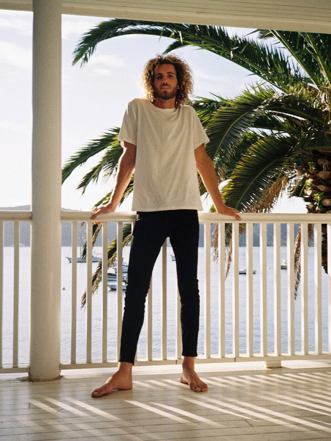 A barefoot person with curly hair, dressed in a white t-shirt and Rollas Stinger Black Gold super stretch skinny jeans, stands on a porch overlooking water and palm trees on a sunny day.