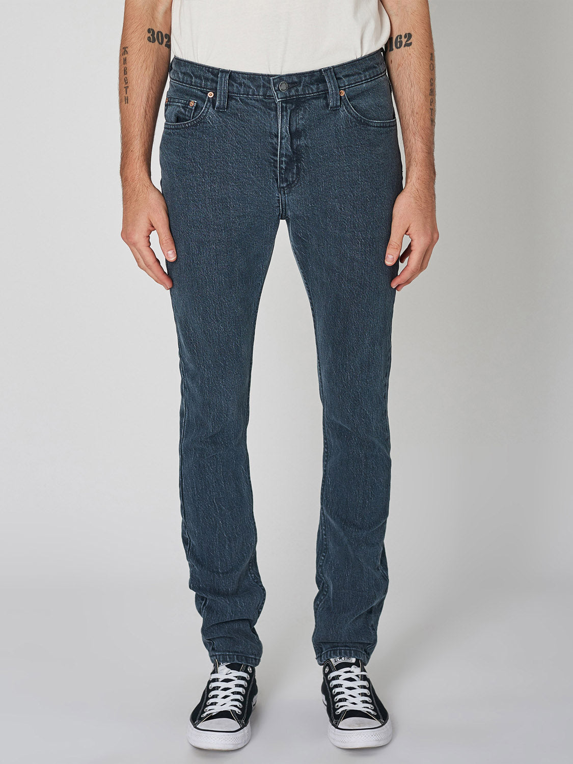 Wearing Rollas Stinger Twisties Inky Black jeans, a white T-shirt, and black-and-white sneakers, a person stands against a plain background with relaxed arms, shown from the shoulders down.