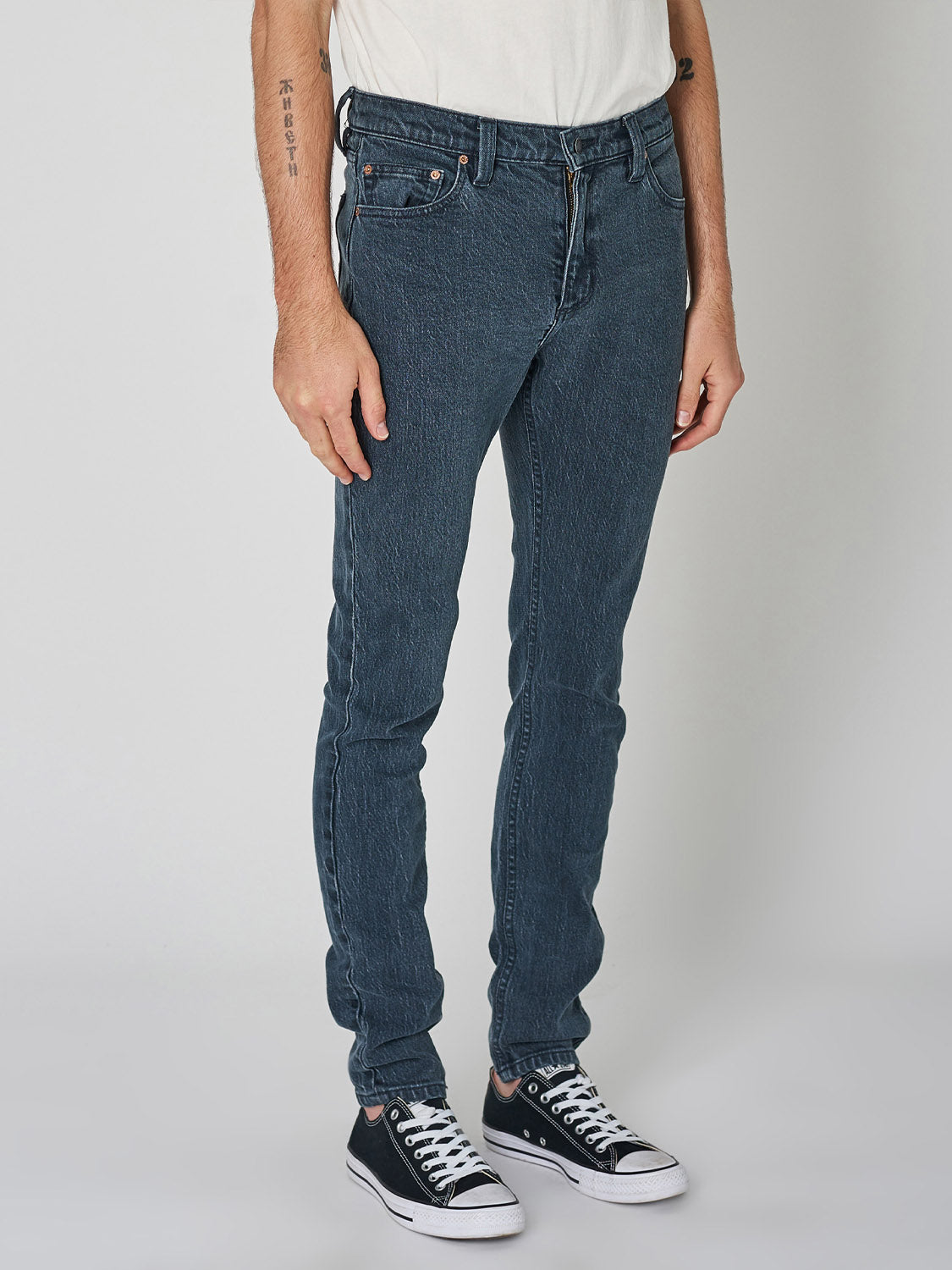 Only the lower half of a person wearing Rollas Stinger Twisties Inky Black skinny jeans, a white T-shirt, and black Converse sneakers is shown standing against a plain light background.