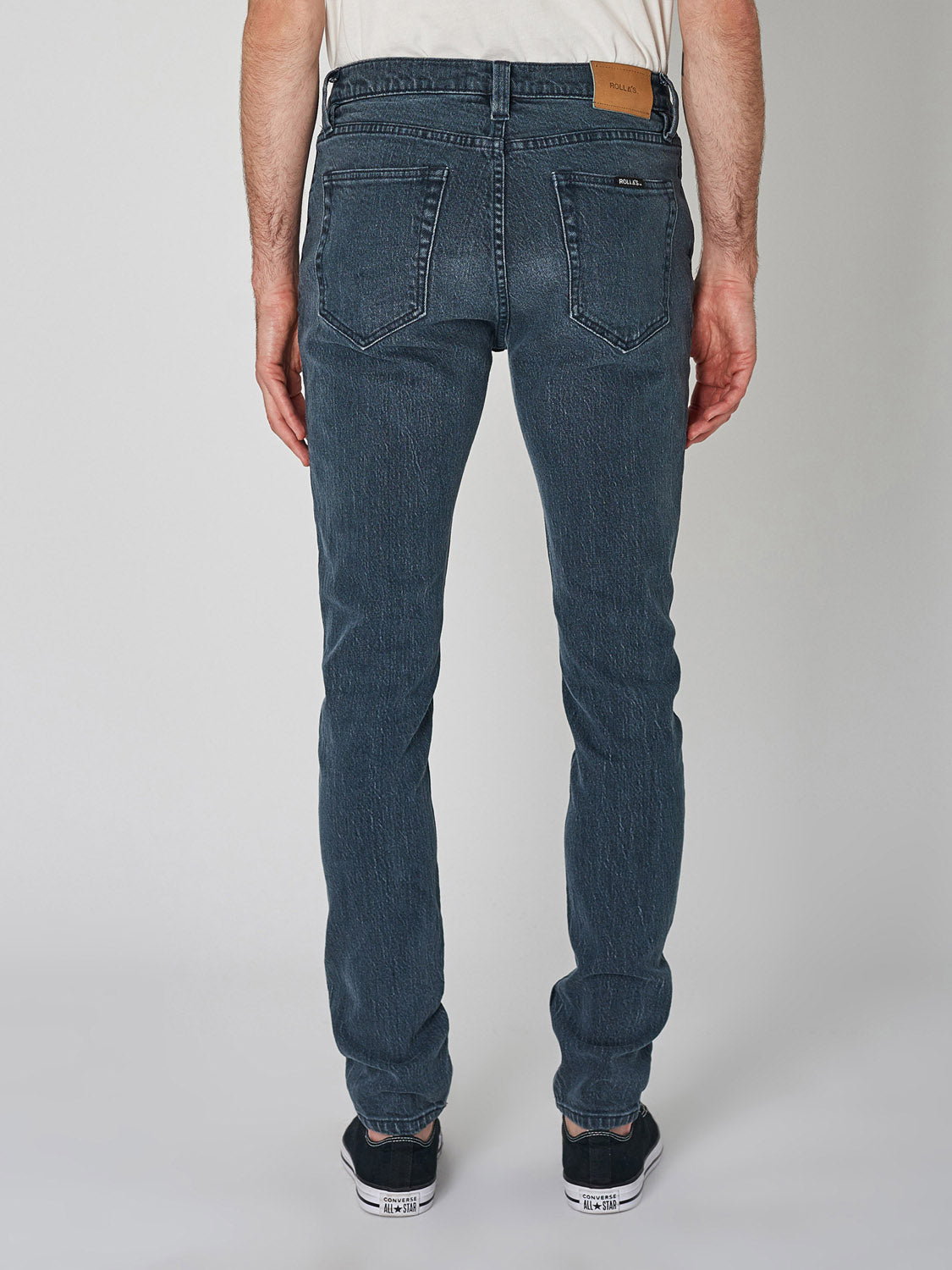 A person stands facing away, wearing a white shirt and Rollas Stinger Twisties Inky Black skinny jeans with elastic denim, back pockets, and a tan waistband patch, paired with black sneakers, against a plain light background.