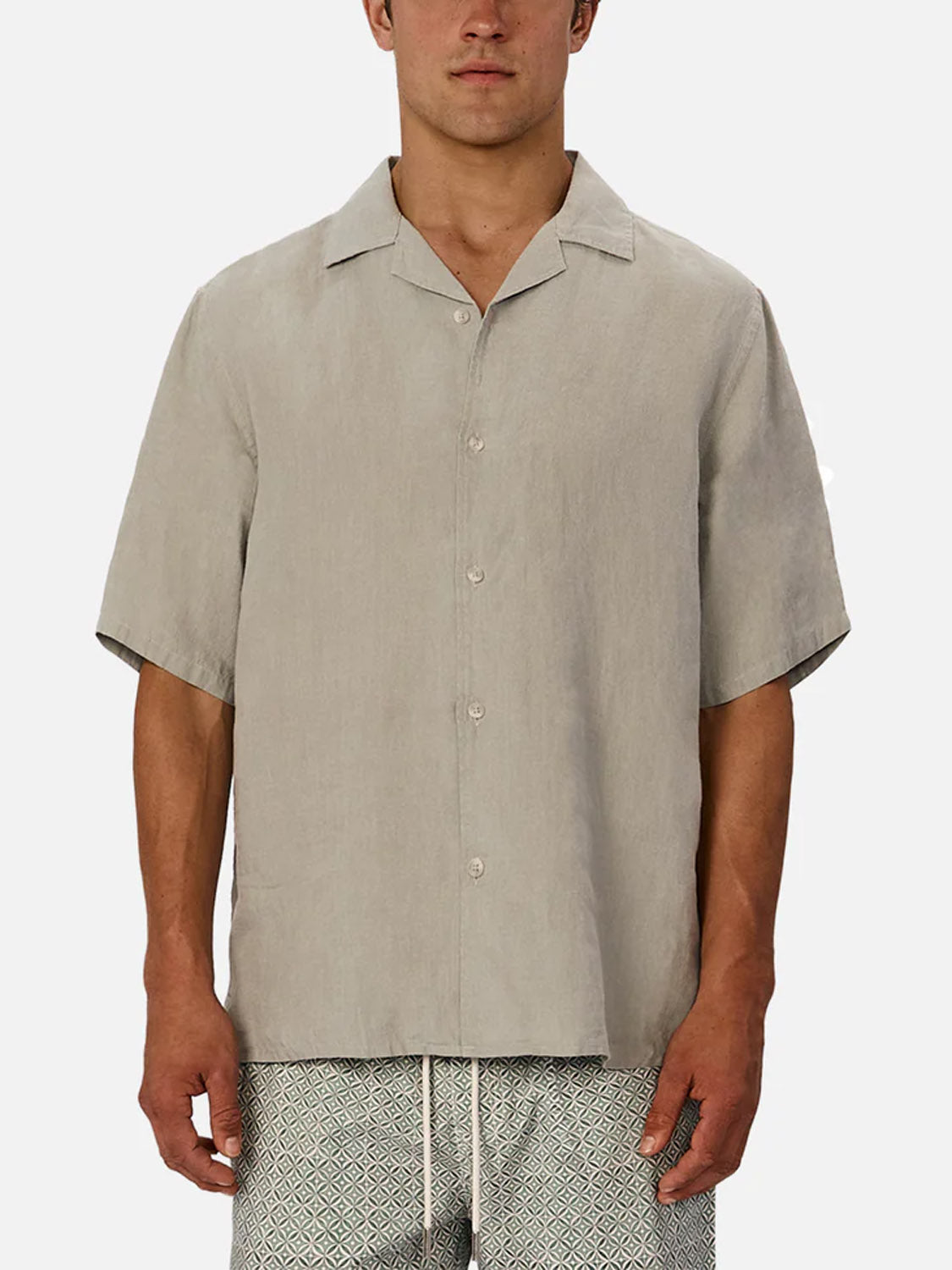 A man models Industrie’s The St Martins Short Sleeve Shirt in light beige with a collar, styled with patterned shorts featuring a white drawstring, standing against a plain white background for a smart casual look.