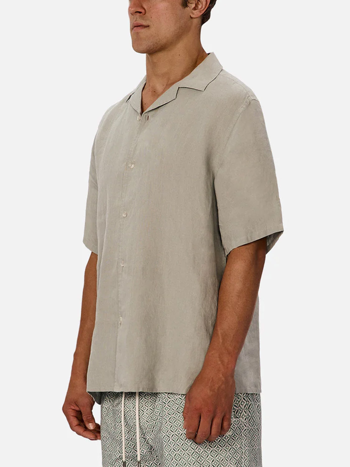 Wearing Industrie’s The St Martins Short Sleeve Shirt in light beige with patterned drawstring shorts, a man stands side-on against a plain white background, showcasing a smart casual look.