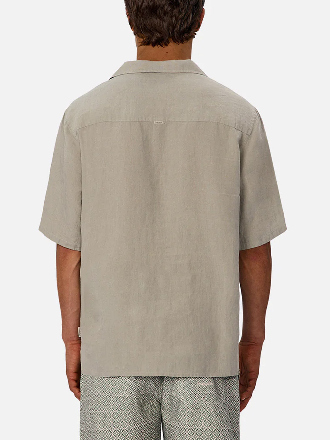 A man is seen from behind wearing Industrie’s The St Martins Short Sleeve Shirt in light beige, paired with geometric patterned shorts, standing against a plain light background for a smart casual look.