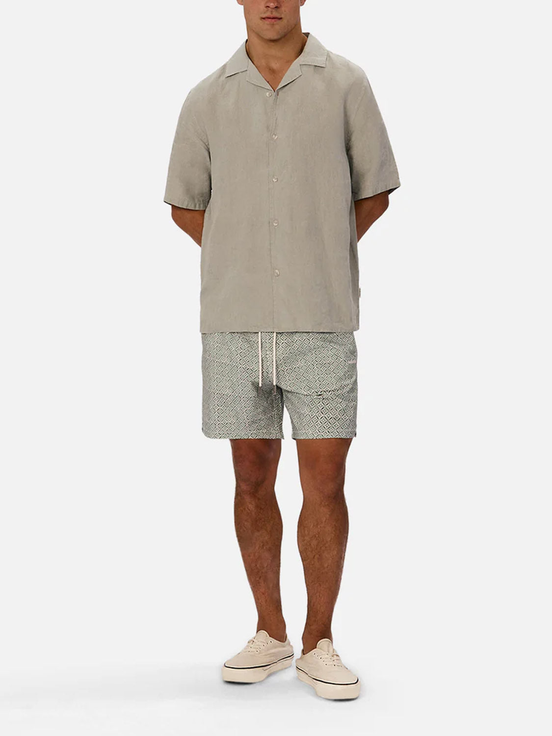 A man faces forward with his hands behind his back, wearing Industrie’s The St Martins Short Sleeve Shirt in light gray, patterned shorts, and beige sneakers for a smart-casual look against a plain white background.