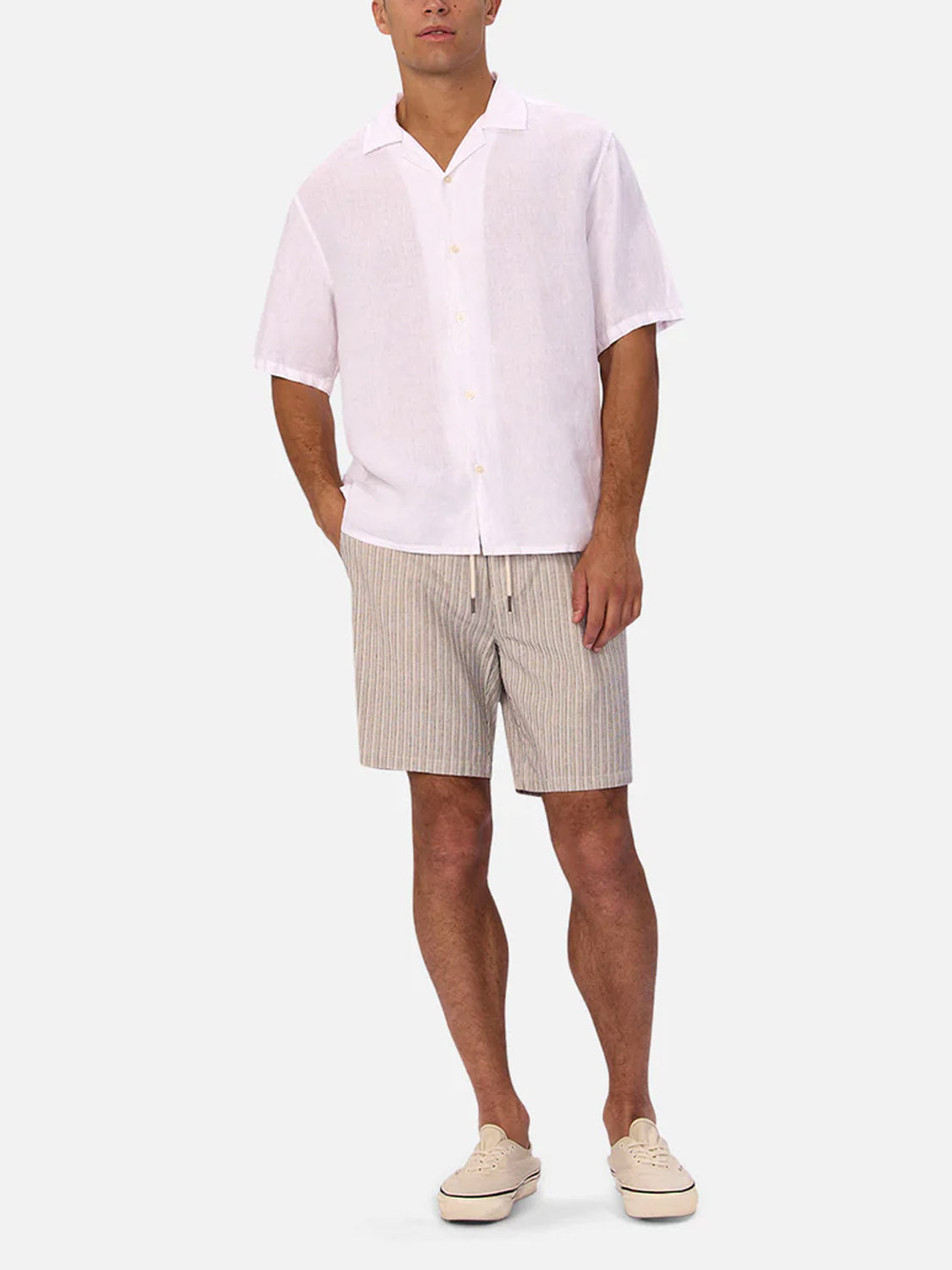 A man in a smart casual look wears The St Martins Short Sleeve Shirt by Industrie with beige and white striped shorts and beige slip-on shoes, standing with hands in pockets against a plain light background.