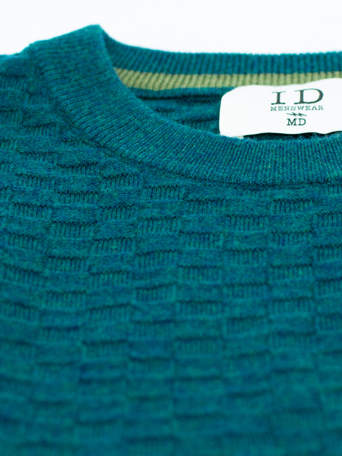 Close-up of the Stockholm Merino Wool Sweater by ID Menswear in textured teal, featuring a ribbed neckline and a white label with ID Menswear in serif font. The lightweight fabric displays a subtle checkered pattern.