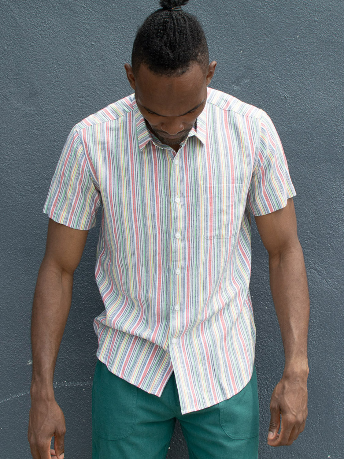 A man with a topknot stands against a gray wall, looking down. He wears the regular fit Streak Cotton Shirt by ID Menswear, featuring lightweight, breathable cotton with vertical pastel stripes, paired with teal pants and his arms at his sides.