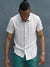 A man with a topknot stands against a gray wall, looking down. He wears the regular fit Streak Cotton Shirt by ID Menswear, featuring lightweight, breathable cotton with vertical pastel stripes, paired with teal pants and his arms at his sides.