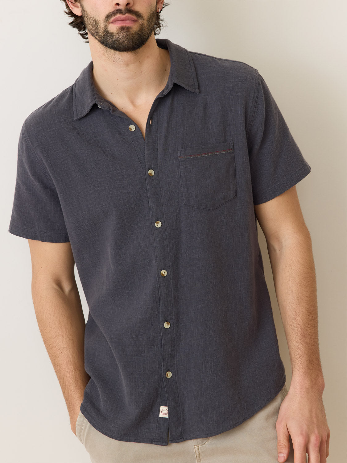 A person wears Marine Layers Stretch Selvage Short Sleeve Shirt in dark gray, featuring a chest pocket and added stretch, paired with beige pants. They stand against an off-white wall, showcasing a relaxed yet refined look that captures the essence of Marine Layers timeless style.