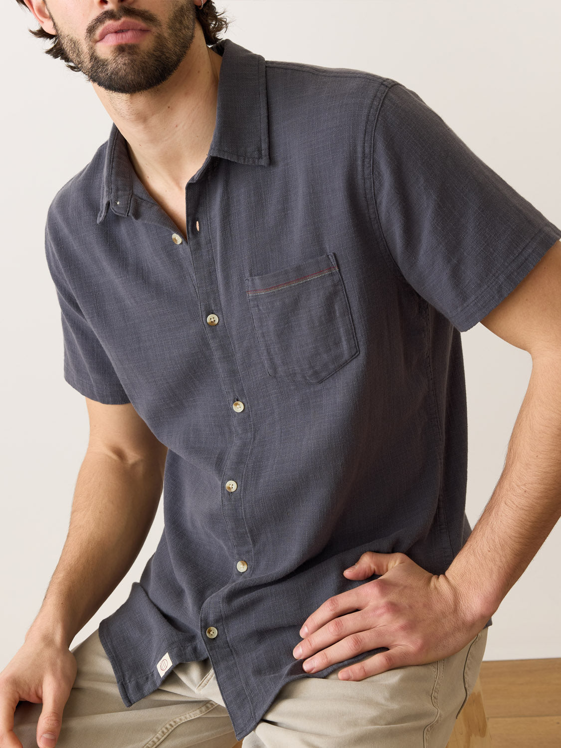 A man sits on a wooden surface wearing the Marine Layer Stretch Selvage Short Sleeve Shirt, featuring a chest pocket. Perfectly paired with light-colored pants against a plain, neutral backdrop for an ideal minimalist setting.