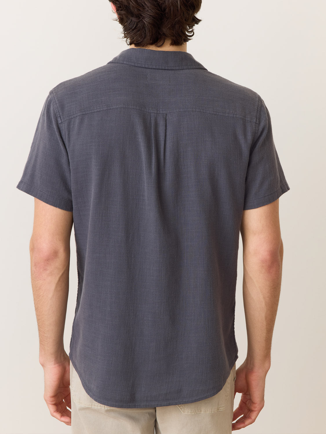 A person is seen from the back in a Marine Layer Stretch Selvage Short Sleeve Shirt, dark gray and featuring added stretch, paired with light pants. The shirts simple collar and straight hem contrast against the plain, light background.