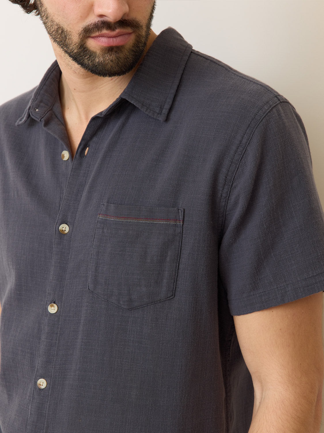 A bearded person wears the Marine Layer Stretch Selvage Short Sleeve Shirt in charcoal, featuring a chest pocket and added stretch. The photo highlights the shirts texture and button details, capturing its classic appeal.