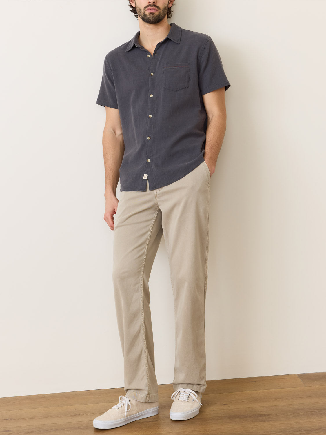 A man in a Marine Layer Stretch Selvage Short Sleeve Shirt, paired with beige stretch pants and white sneakers, stands against a light wall on a wooden floor. With hands in pockets, his look is enhanced by his beard and short hair.