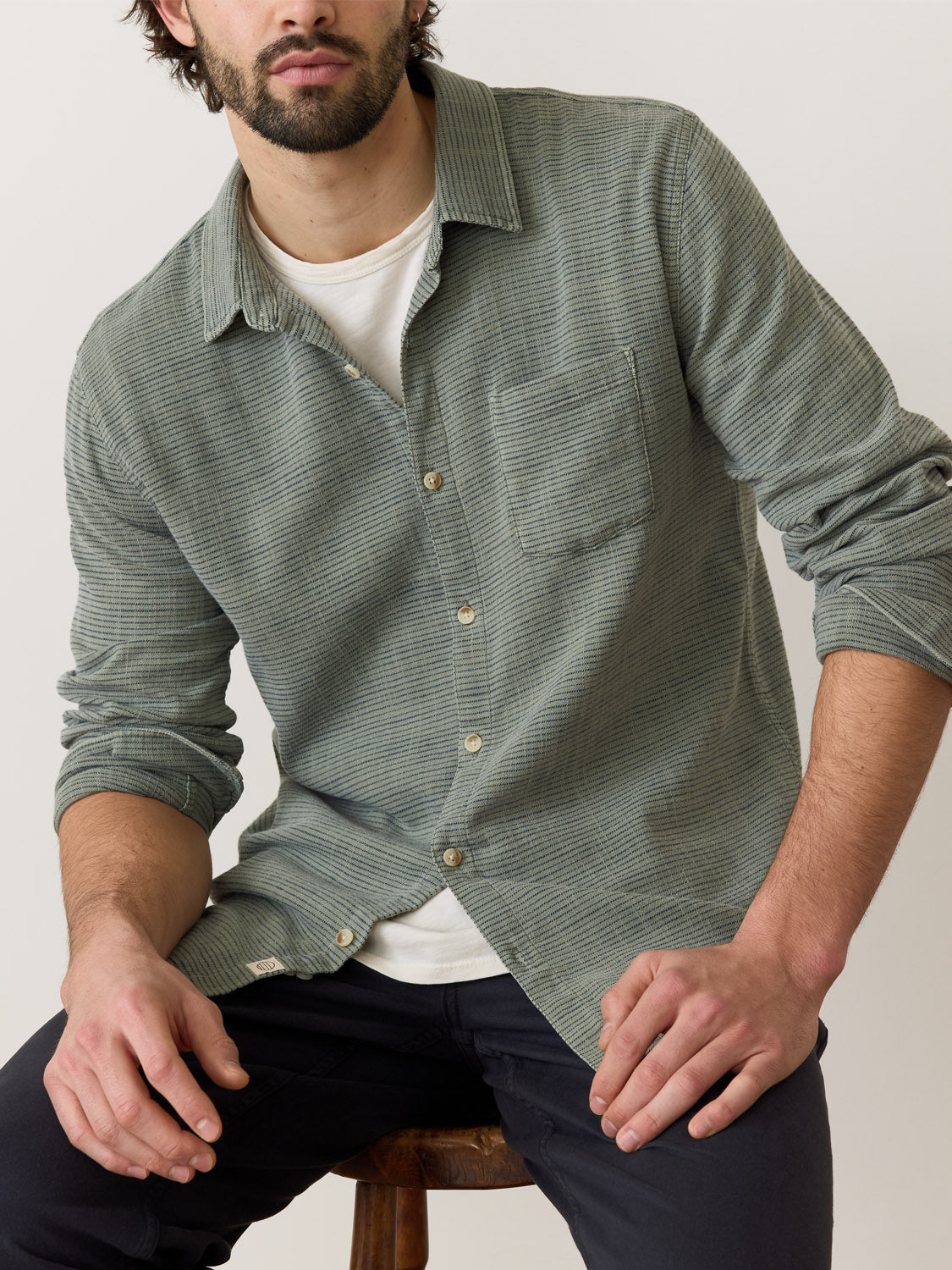 A man sits on a stool wearing the Marine Layer Stretch Selvage Long Sleeve Shirt. The green textured design complements his white top and dark pants as he leans forward, hands on thighs, against a plain, light backdrop.