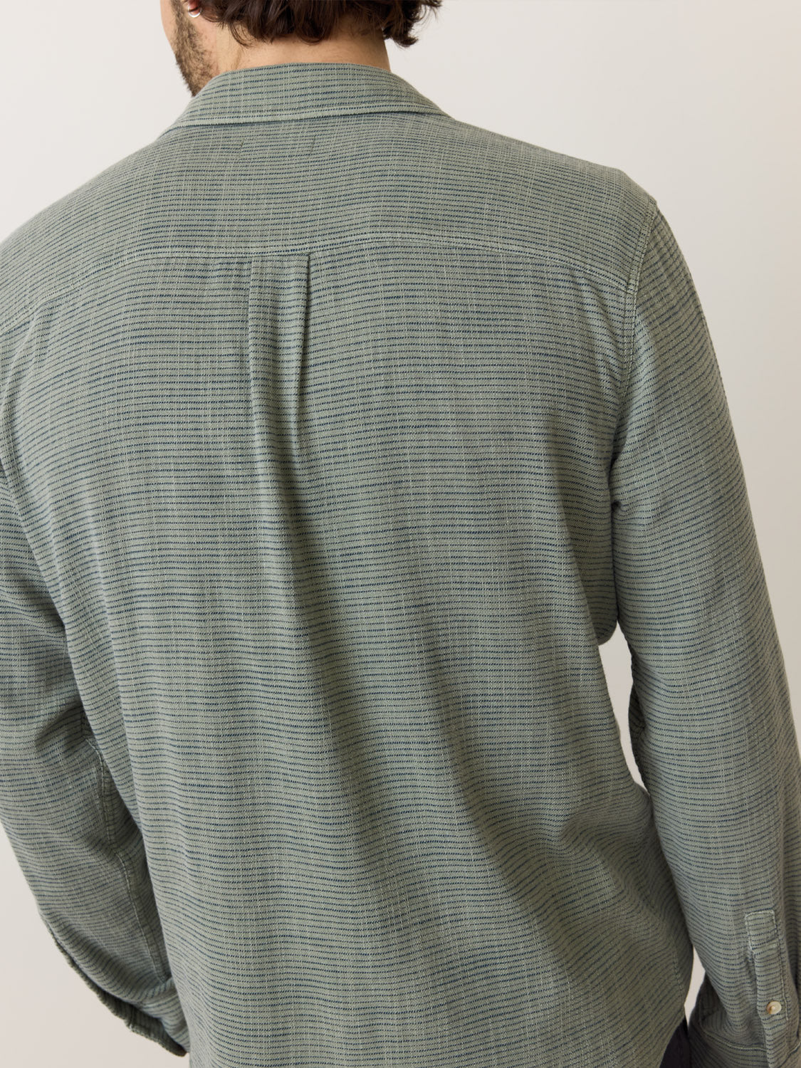 A person is seen from behind wearing a Marine Layer Stretch Selvage Long Sleeve Shirt. This gray-green checkered Marine Layer shirt has long sleeves, a button-down design, and offers a slightly textured pattern for added stretch against an off-white backdrop.