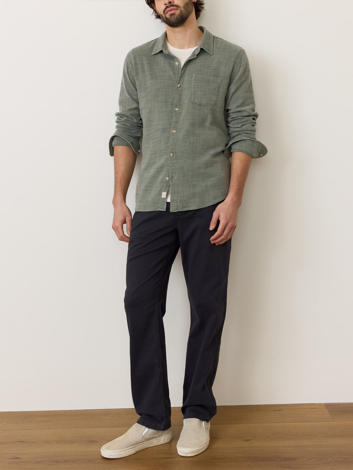 A man stands against a white wall wearing a Marine Layer Stretch Selvage Long Sleeve Shirt, paired with dark pants and beige slip-on shoes. His hands rest at his sides, blending into the rooms light wooden floor.