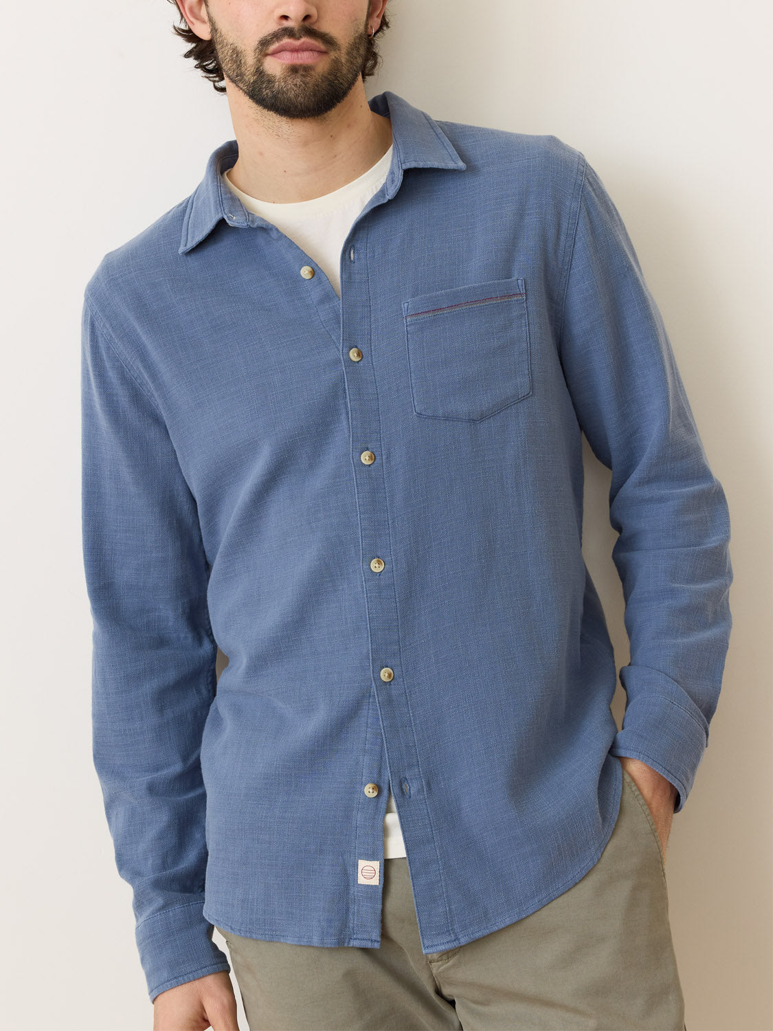 Dressed in a Marine Layer Stretch Selvage Long Sleeve Shirt with added stretch over a white t-shirt, a person stands against a light background. The shirt has a left chest pocket, and they are wearing beige pants to complete the look.