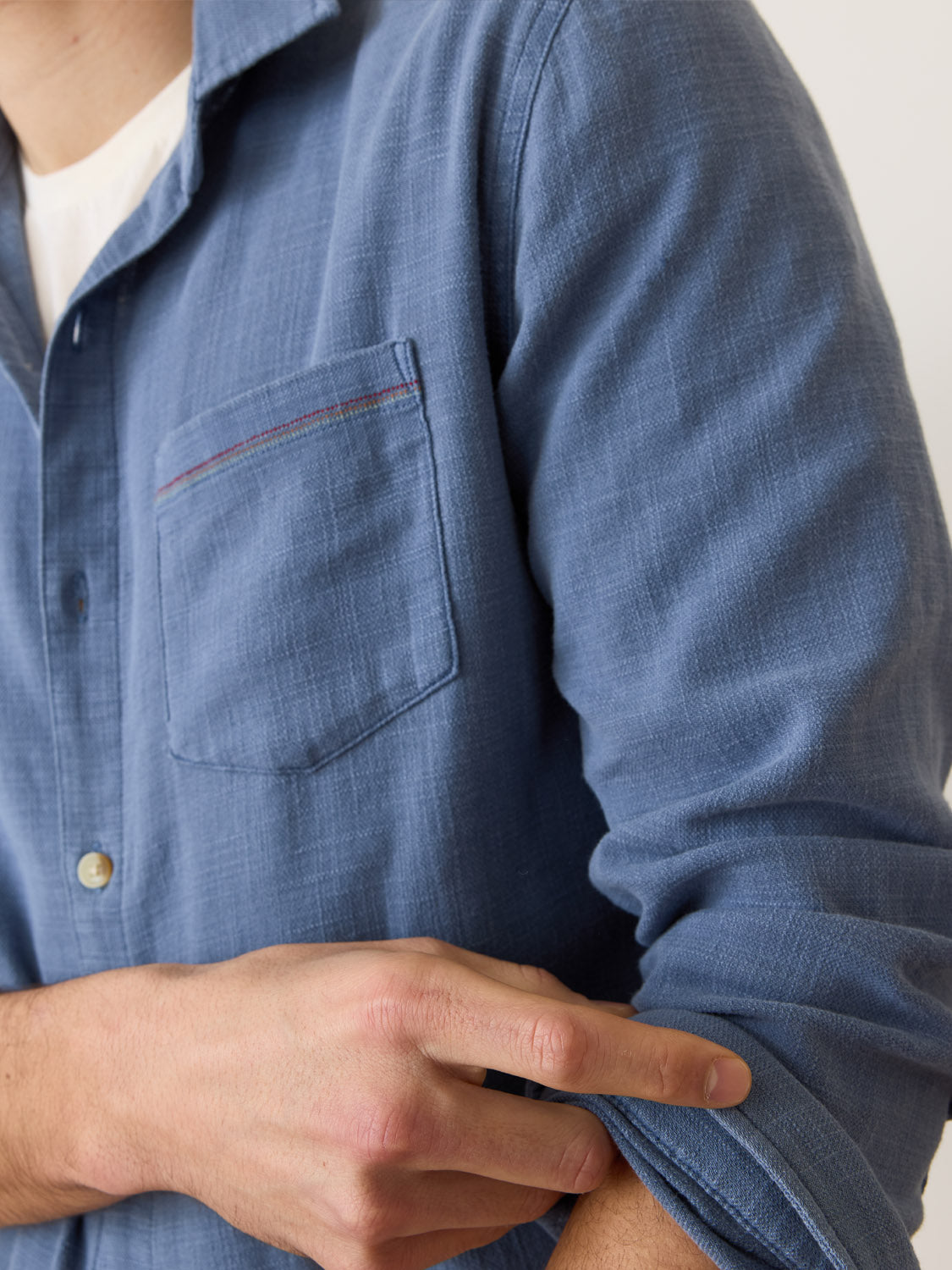 Wearing a Marine Layer Stretch Selvage Long Sleeve Shirt in marine blue, someone adjusts their rolled-up sleeve. The stylish shirt, layered over a white T-shirt, stands out against the plain light background.