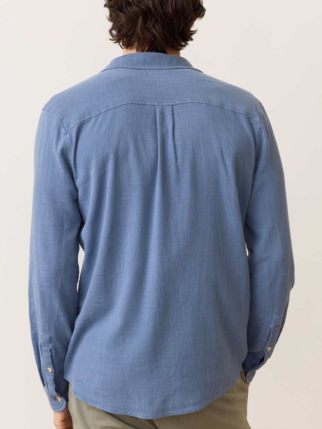 A person is seen from the back wearing a Marine Layer Stretch Selvage Long Sleeve Shirt in blue, featuring buttoned cuffs and a casual fit with added stretch. They pair it with beige pants, set against a simple, light-colored background.