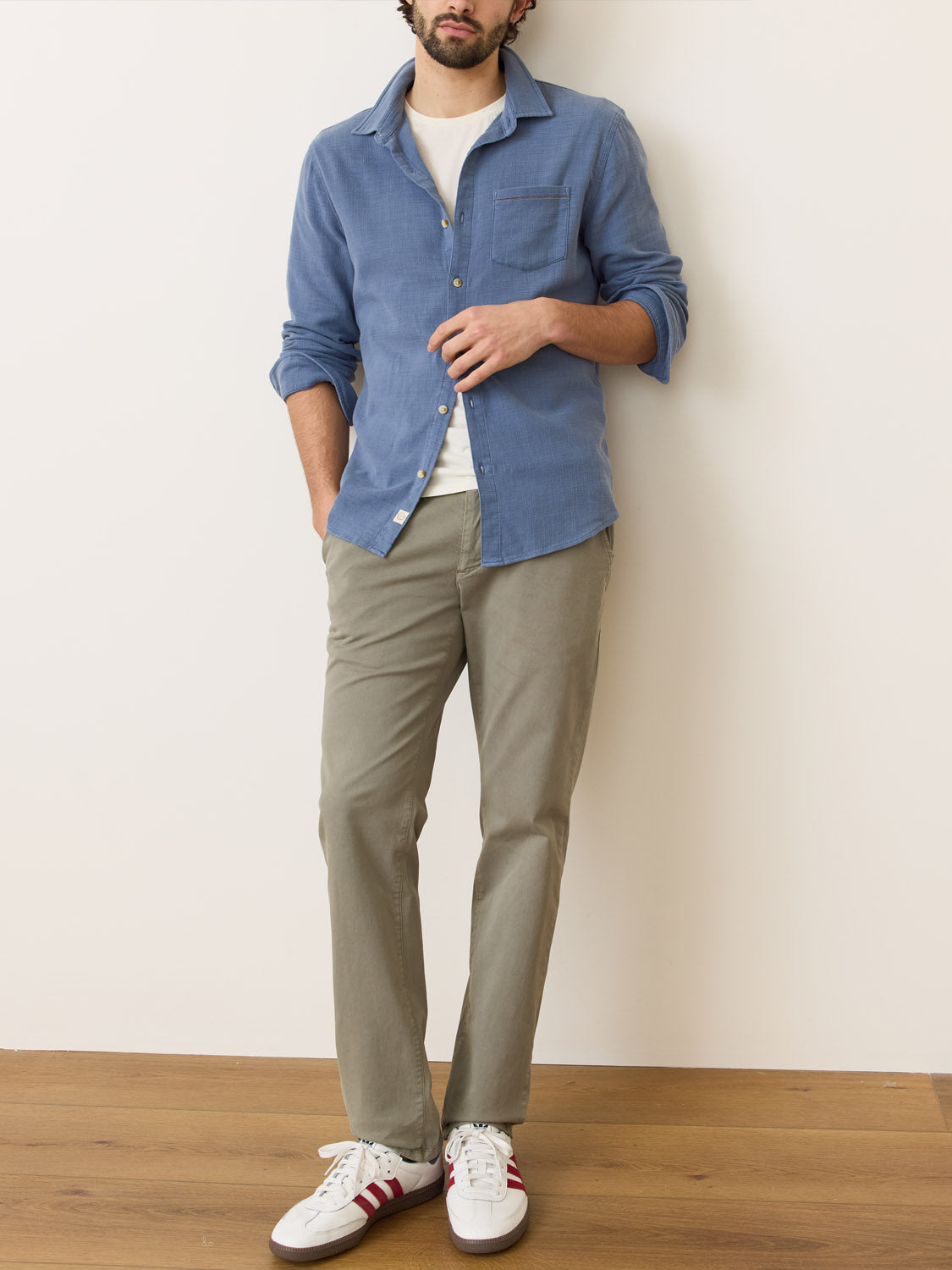 A man casually leans against a white wall and wooden floor, exuding effortless style in a Marine Layer Stretch Selvage Long Sleeve Shirt over a white tee, paired with khaki pants and red-striped white sneakers.