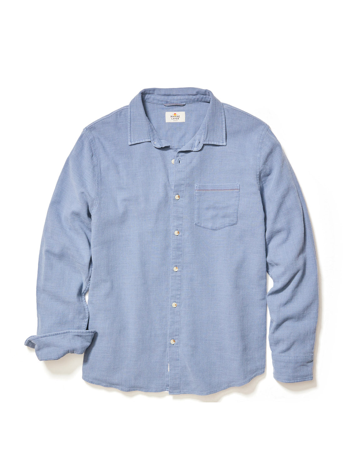 The Marine Layer Stretch Selvage Long Sleeve Shirt in light blue features textured fabric, a single chest pocket, and stylish selvage detailing on a partially rolled-up sleeve against a plain white backdrop.