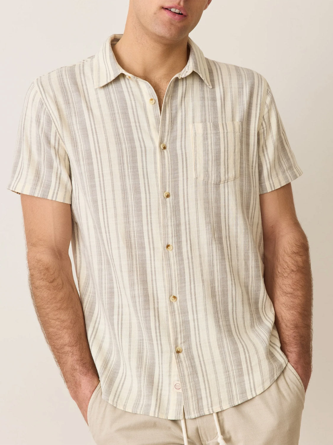A person stands against a plain background wearing the Marine Layer Stretch Selvage Short Sleeve Shirt. This light-colored, vertically striped shirt by Marine Layer has embroidered details and a classic fit with a pocket. The individuals hands are casually tucked into their pockets.