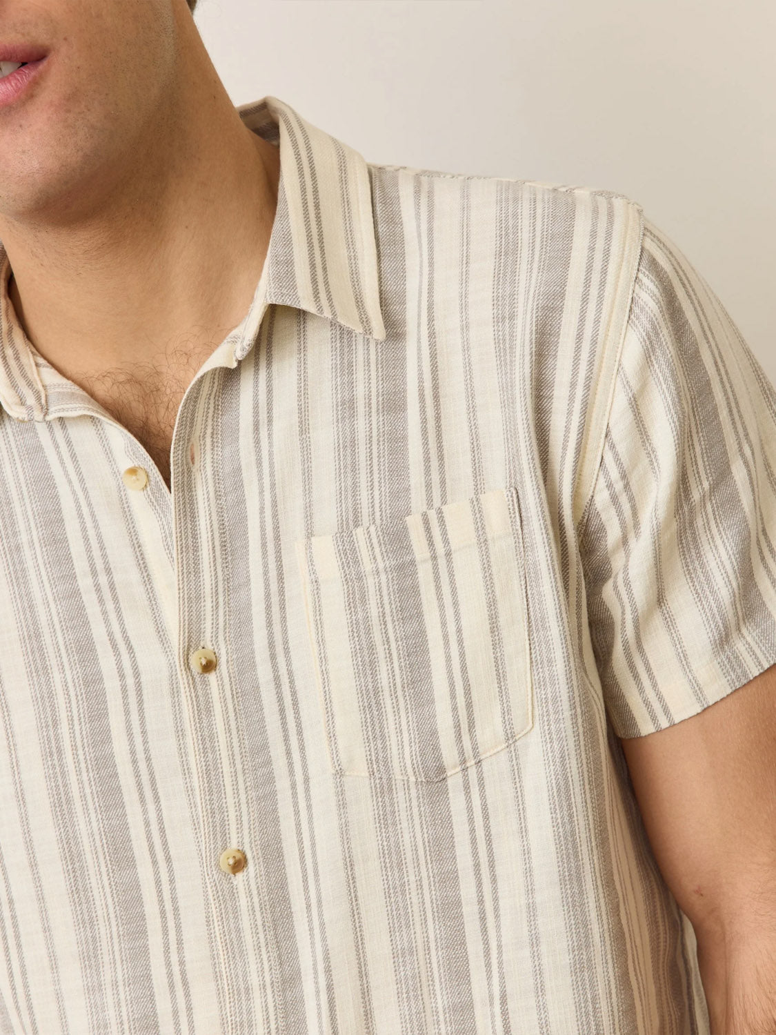 A close-up shows a person in the Marine Layer Stretch Selvage Short Sleeve Shirt by Marine Layer. Its a classic fit with beige and gray stripes, embroidered details, and a chest pocket. The top button is undone to show part of the neck against a neutral background, highlighting its refined style.