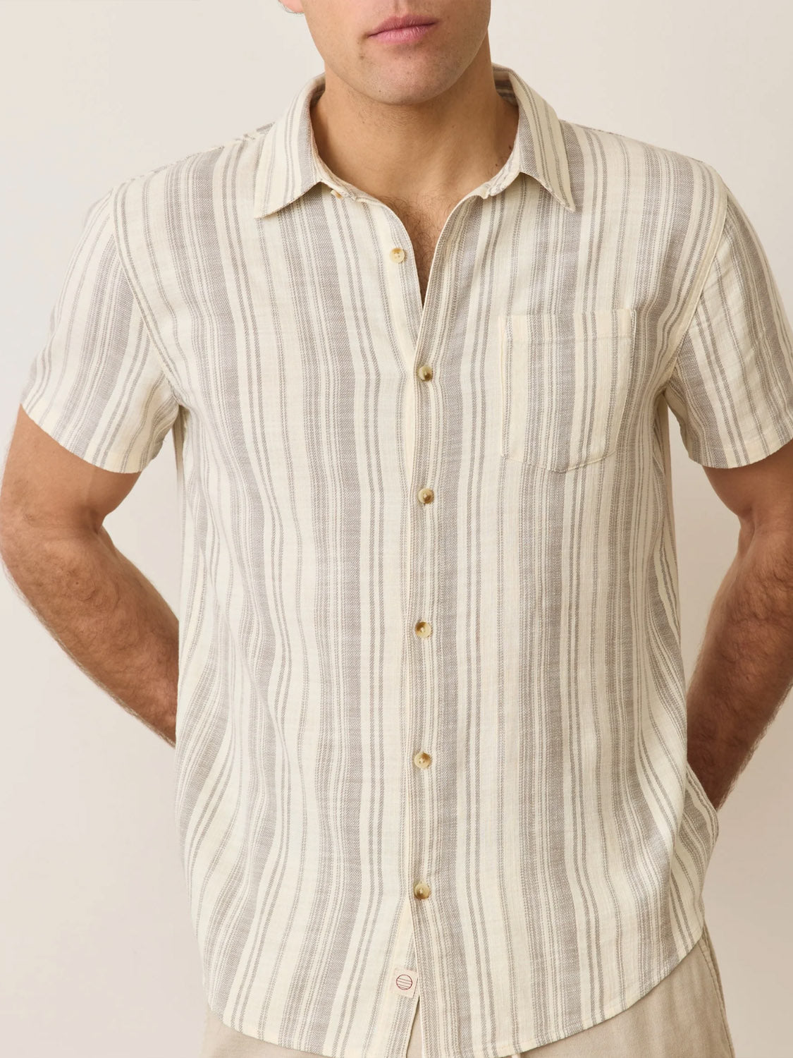 A man stands with his arms behind his back, wearing the elegant Marine Layer Stretch Selvage Short Sleeve Shirt, a classic fit button-up with beige and gray stripes and an embroidered chest pocket. The scene exudes understated style against an off-white backdrop.