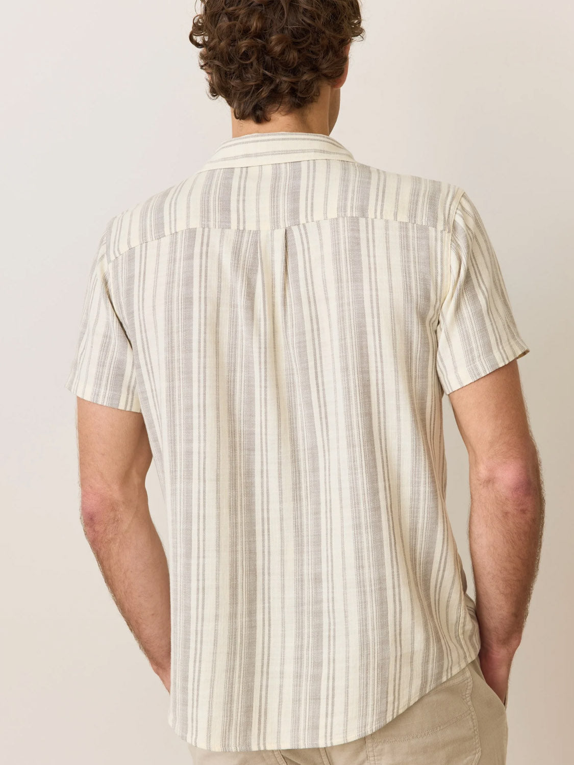 A person with curly hair wears the Marine Layer Stretch Selvage Short Sleeve Shirt featuring vertical stripes and embroidered details. The classic fit, light-colored shirt is paired with jeans. They face away, revealing the back against a neutral background.