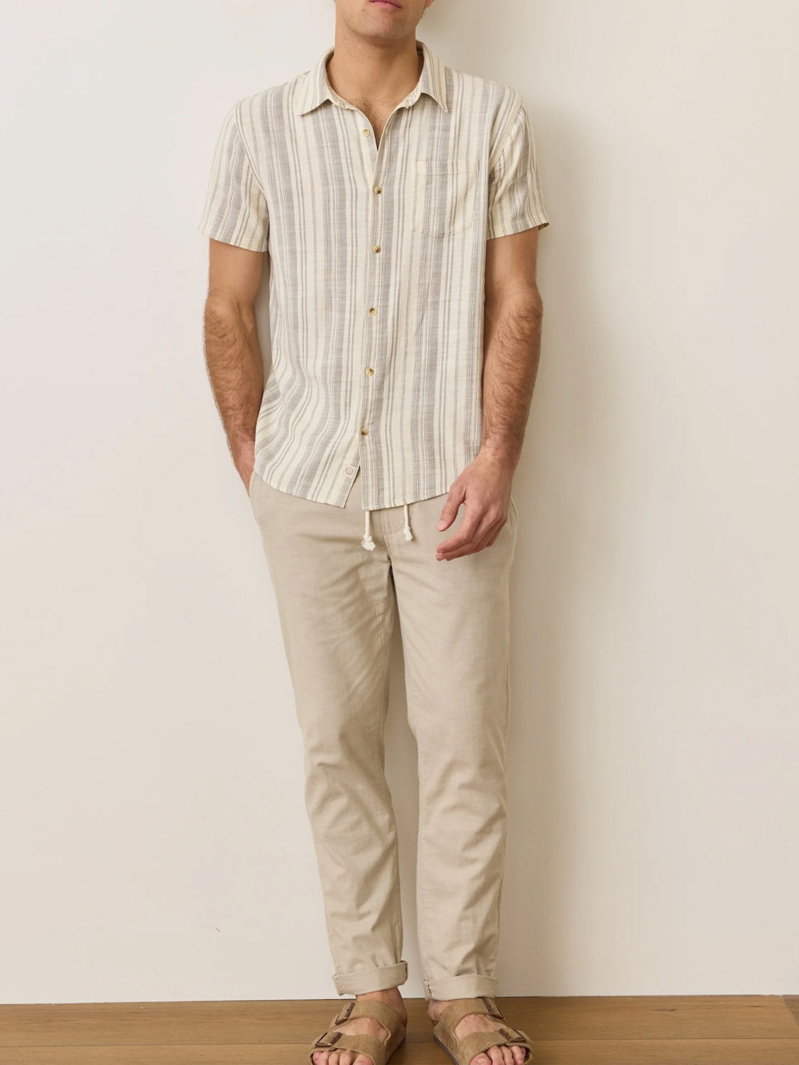 A person wearing the Marine Layer Stretch Selvage Short Sleeve Shirt in beige and white stripes, paired with beige drawstring pants and brown sandals, stands against a plain light-colored wall on a wooden floor.
