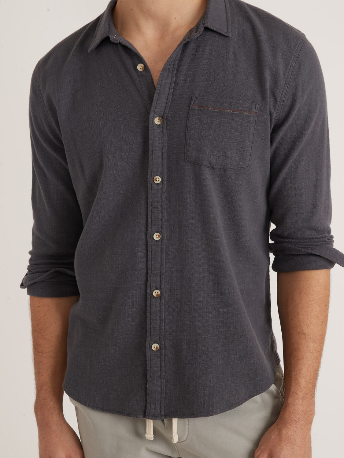 A person is shown from shoulders to waist wearing a Marine Layer Stretch Selvage Long Sleeve Shirt in charcoal gray, featuring a left chest pocket and added stretch. The stylish shirt is paired with light-colored pants, creating a classic yet comfortable look.