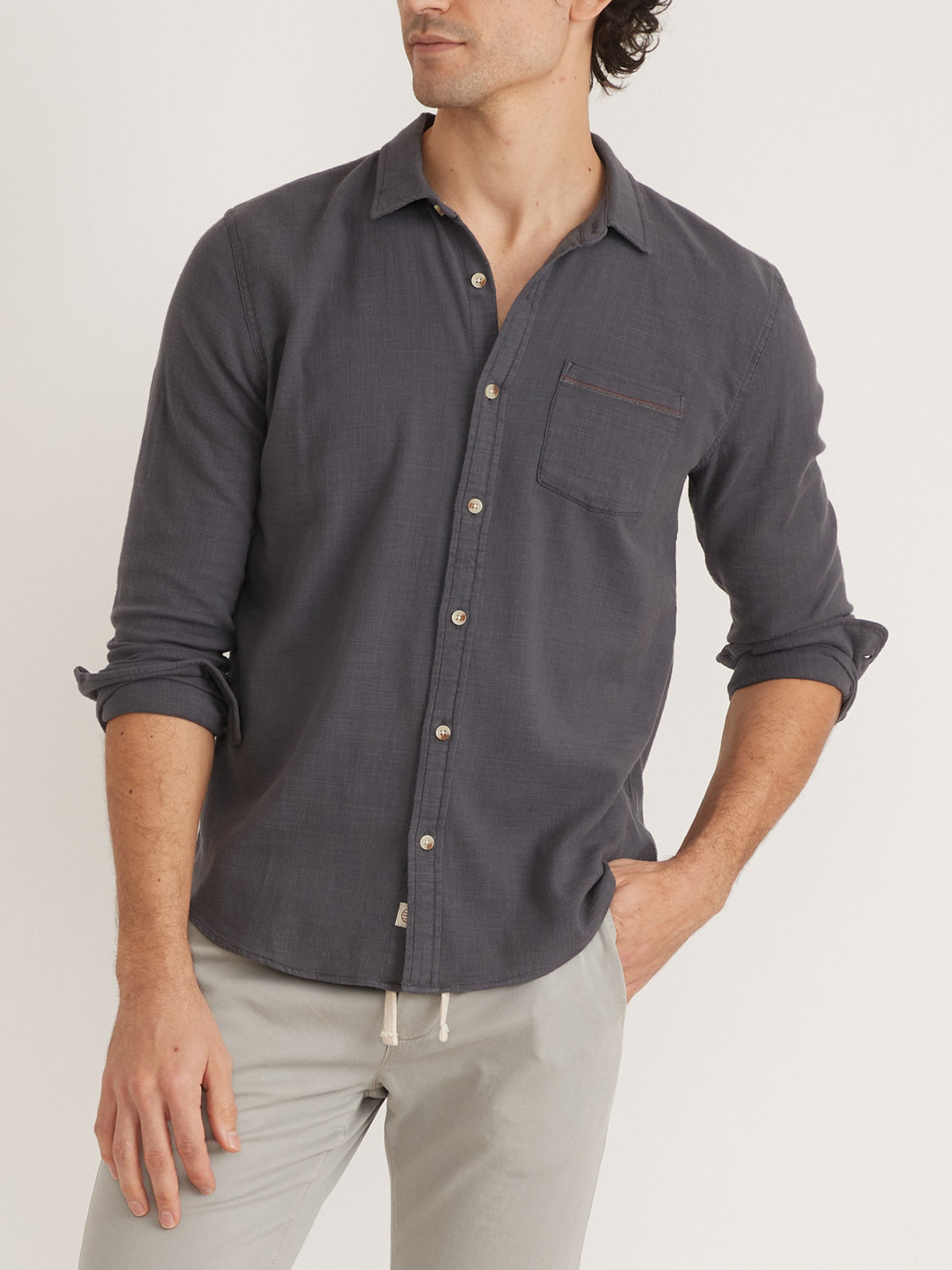 A person radiates relaxed Marine Layer style, wearing a Marine Layer Stretch Selvage Long Sleeve Shirt in dark gray with rolled sleeves and light gray pants, standing against a plain background with one hand in their pocket.