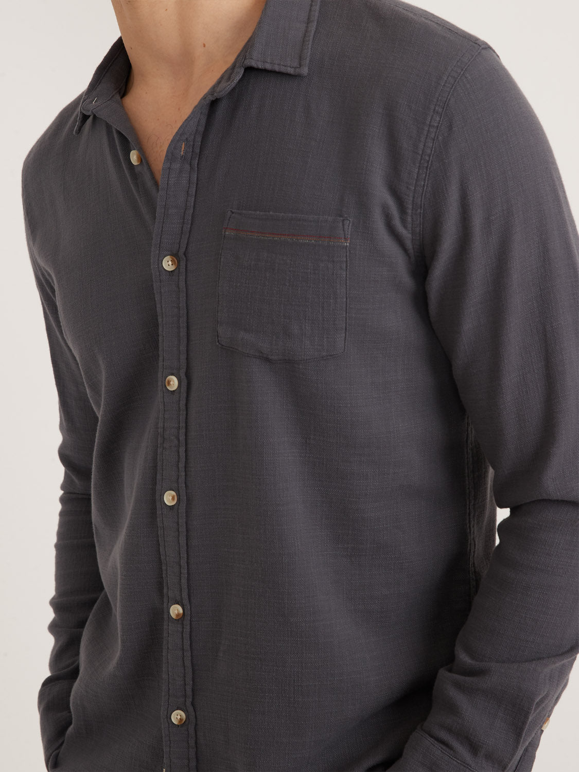 Someone is wearing a dark gray Marine Layer Stretch Selvage Long Sleeve Shirt with a collar, chest pocket, and light brown buttons. The shirt incorporates added stretch for comfort, enhancing its casual look. The persons head isnt visible in the image.