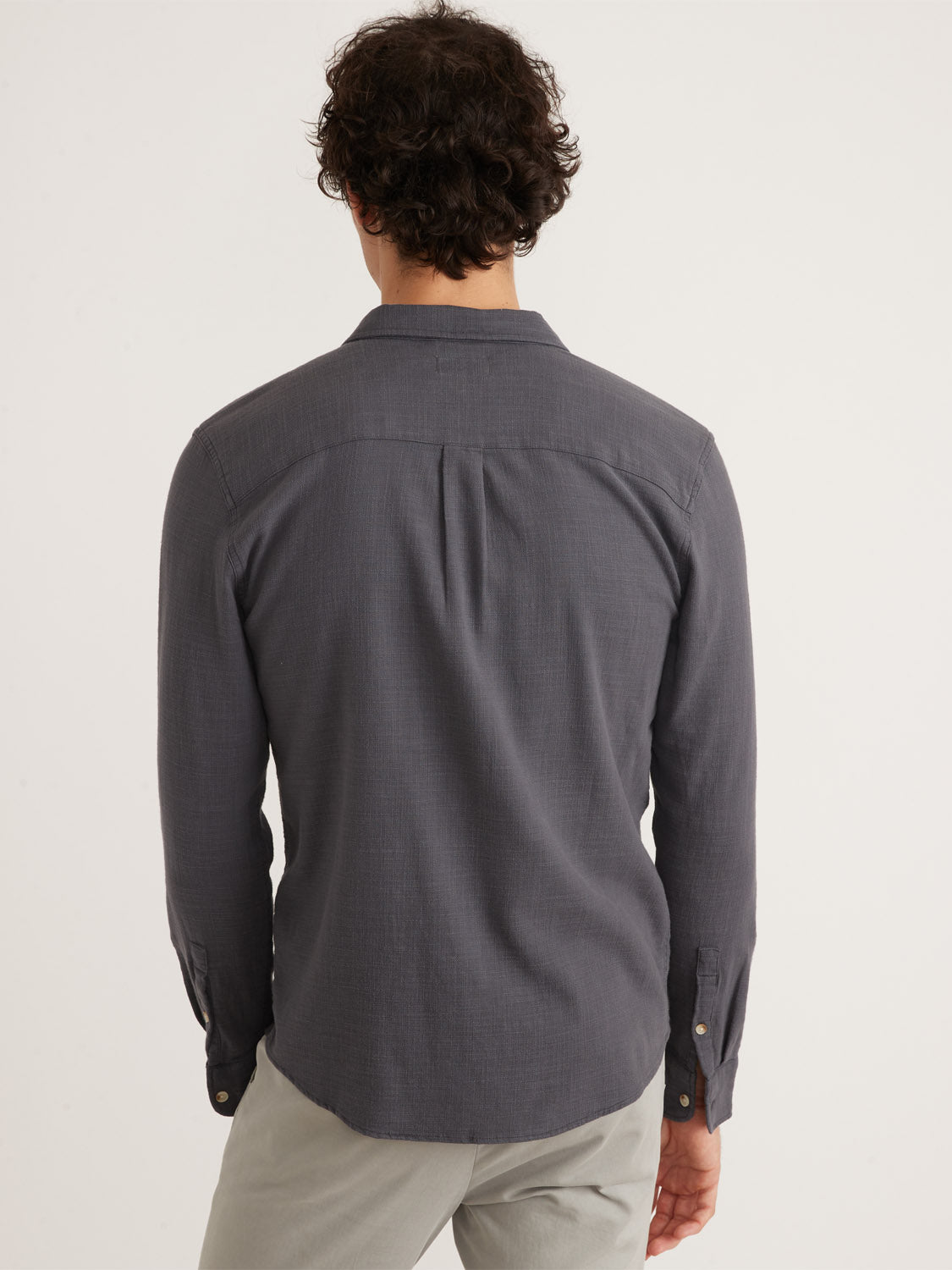 A person with curly hair wears a Marine Layer Stretch Selvage Long Sleeve Shirt and light pants, standing against a plain background.