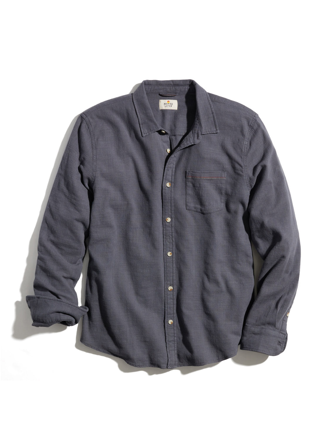 The Marine Layer Stretch Selvage Long Sleeve Shirt in dark gray has a subtle texture and added stretch. It features a collar, long sleeves, and a left-side chest pocket, laid flat against a white background.