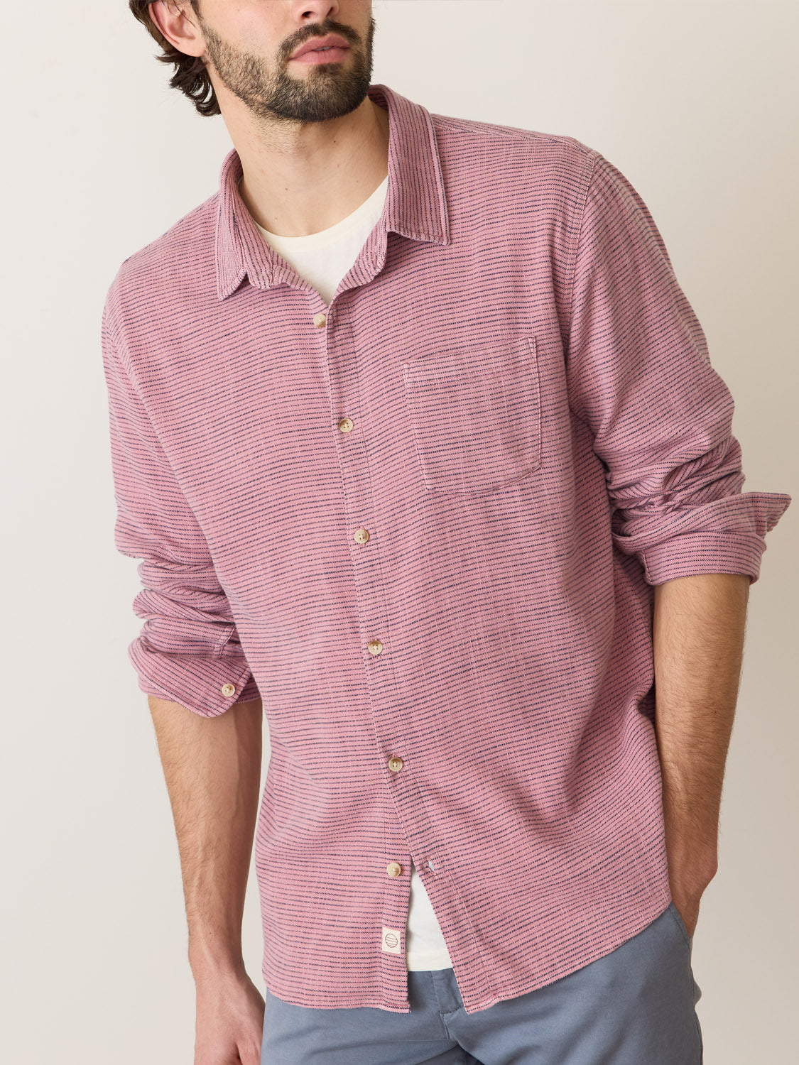 A man wears a Marine Layer Stretch Selvage Long Sleeve Shirt in red stripes over a white T-shirt, with its sleeves slightly rolled. He pairs it with gray pants against a pure white background.